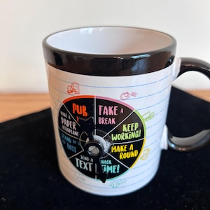 May include: White ceramic mug with a black rim and handle. The mug features a colorful wheel design with phrases like "Take a Break" and "Text Me!" against a notebook paper background.