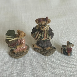 May include: Three miniature figurines featuring two anthropomorphic bears and a small mouse. One bear wears a pink dress and carries a stack of books, the other wears a dark coat and glasses, and the mouse holds a small sign labeled CAT.