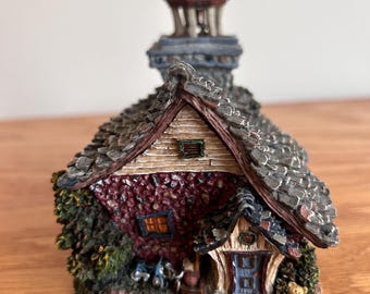 May include: A detailed miniature house figurine with a red brick exterior, gray shingled roof, and a small tower. The house features a small blue door, a wooden porch, and decorative landscaping with tiny figures and a teddy bear.