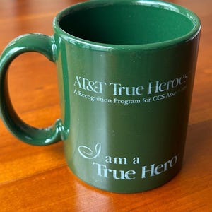 May include: A green ceramic mug with a curved handle. The mug features the text "AT&T True Heroes" and "I am a True Hero" in white lettering. The mug is cylindrical and appears to be empty.