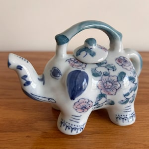 May include: A white ceramic teapot in the shape of an elephant, decorated with blue and pink floral patterns. The teapot has a curved handle and a small lid. The spout is on the elephant's trunk.