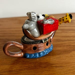 May include: A ceramic Noah's Ark-themed trinket box. The ark is a brown teacup with a blue wave design at the base. Inside are gray elephants, a white bear, a giraffe, and a red box. The giraffe's head extends from the cup.