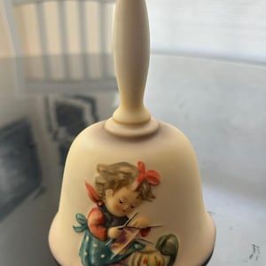 May include: A cream-colored ceramic bell with a long handle, featuring a painted illustration of a person with brown hair and a red bow, wearing a blue and red outfit while knitting with needles and yarn, with a small green object nearby.