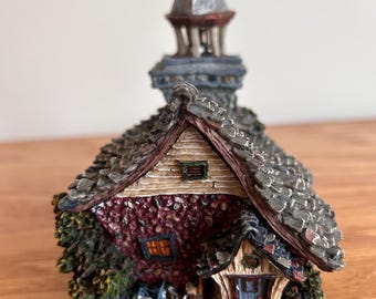 May include: A miniature house figurine featuring a dark red stone-textured exterior, a gray shingled roof, and a central tower with a dome. The structure includes a small blue door, green foliage accents, and a tiny teddy bear near the entrance.
