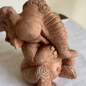 May include: A terracotta elephant sculpture in a seated position, with its trunk curled and its ears covering its eyes. The sculpture has detailed textures and a natural, earthy color, showcasing the artist's craftsmanship.