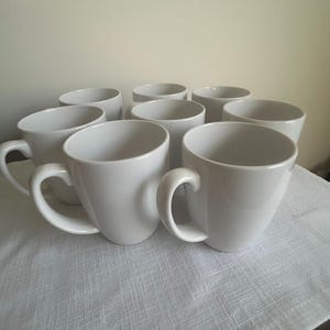 May include: A set of eight white ceramic coffee mugs. Each mug has a rounded body, a curved handle, and a smooth, glossy finish. The mugs are arranged on a white textured tablecloth, perfect for a kitchen or dining setting.