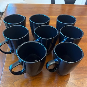 May include: Eight black ceramic mugs with curved handles are arranged on a wooden surface. The mugs have a glossy finish and a simple, modern design. The mugs are empty and ready for use.