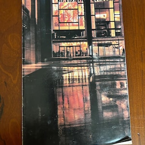 May include: A book cover featuring the Metropolitan Opera. The image shows a building with large windows, reflecting the lights and colors of the interior. The cover has a dark, moody aesthetic.