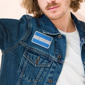 May include: A man wearing a blue denim jacket with a Honduran flag patch on the left side of the chest.