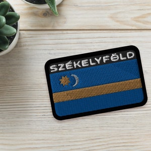Puede incluir: Un parche bordado en blanco y negro con el texto "SZÉKELYFÖLD" en letras blancas. El parche tiene un fondo azul con una franja dorada y una estrella blanca y una luna creciente.