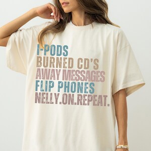May include: Cream-colored t-shirt with the phrases "I-PODS, BURNED CD'S, AWAY MESSAGES, FLIP PHONES, NELLY.ON.REPEAT" printed in various colors. The shirt has short sleeves and a relaxed fit.