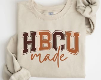 May include: A beige sweatshirt with the text "HBCU made" in brown and orange lettering. The letters "HBCU" are in a collegiate style font. The sleeves are folded and the sweatshirt is on a white background.