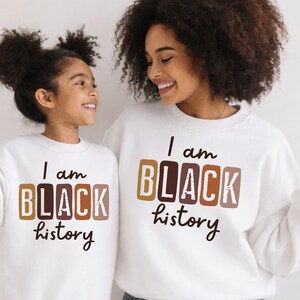 May include: Two people wearing white sweatshirts with the text "I am BLACK history" in brown and tan block letters. The sweatshirts are a matching set, with one worn by a child and the other by an adult.