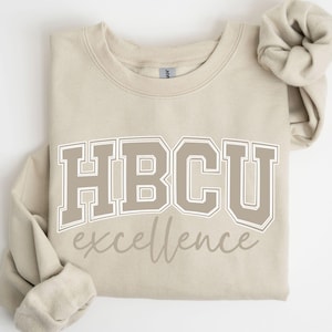 May include: A beige sweatshirt with the text "HBCU excellence" in a collegiate font. The letters are outlined in white with a beige fill. A matching beige scrunchie is next to the sweatshirt.
