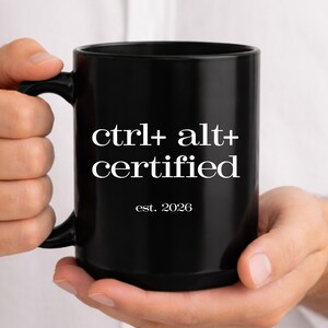 May include: A black ceramic mug with the text "ctrl+alt+certified" in white, with "est. 2026" below. The mug is held by a person.