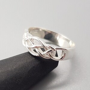 Puede incluir: Un anillo de plata con un diseño de nudo celta. El anillo está sobre una superficie negra.