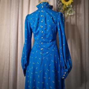 Può includere: Un abito lungo blu con un motivo floreale bianco e un colletto alto. L'abito ha una schiena con bottoni e una vita aderente.