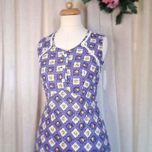 Può includere: Un abito senza maniche con un motivo floreale viola e bianco. L'abito ha un bordo in pizzo bianco attorno al collo e una chiusura con bottone sul davanti.