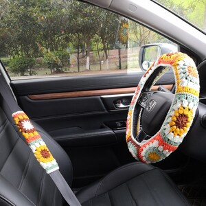 May include: A crocheted steering wheel cover with a floral pattern in shades of yellow, orange, white, and green. The cover is on a black leather steering wheel in a car.
