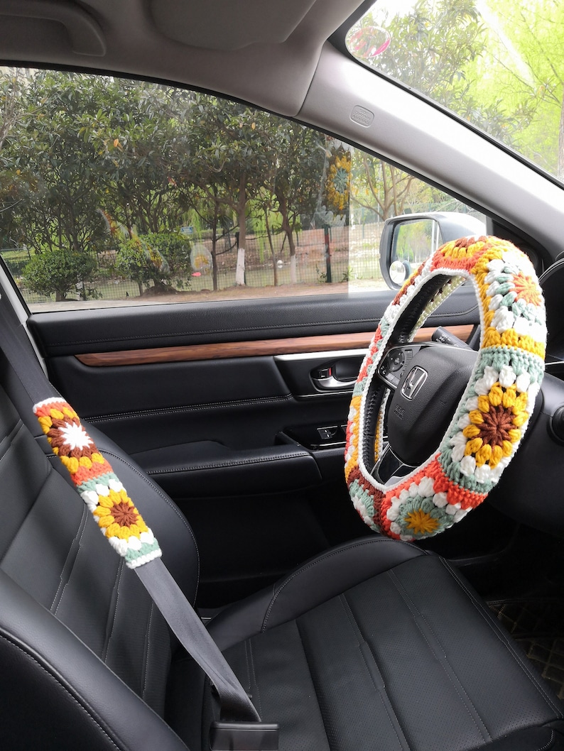 May include: A crocheted steering wheel cover with a floral pattern in shades of yellow, orange, white, and green. The cover is on a black leather steering wheel in a car.