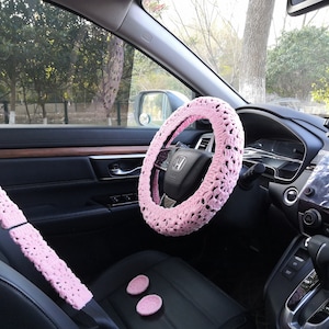 Puede incluir: Cubierta de volante y cubiertas de cintur&oacute;n de seguridad de crochet rosa para el interior de un coche. La cubierta del volante tiene un delicado patr&oacute;n floral.