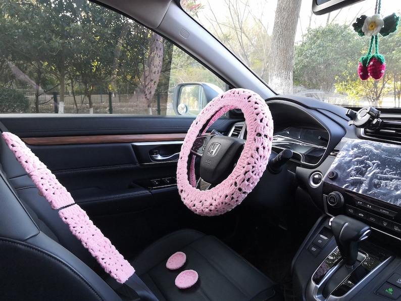 Puede incluir: Cubierta de volante y cubiertas de cintur&oacute;n de seguridad de crochet rosa para el interior de un coche. La cubierta del volante tiene un delicado patr&oacute;n floral.