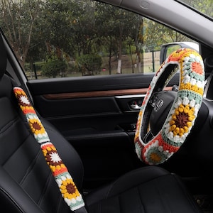 May include: A crocheted steering wheel cover and seatbelt cover in a colorful floral pattern with shades of orange, yellow, green, white, and brown. The cover is made of yarn and has a granny square design.