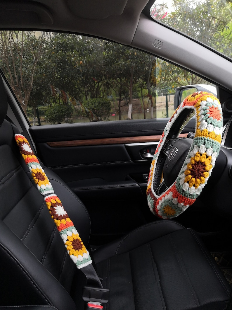 May include: A crocheted steering wheel cover and seatbelt cover in a colorful floral pattern with shades of orange, yellow, green, white, and brown. The cover is made of yarn and has a granny square design.