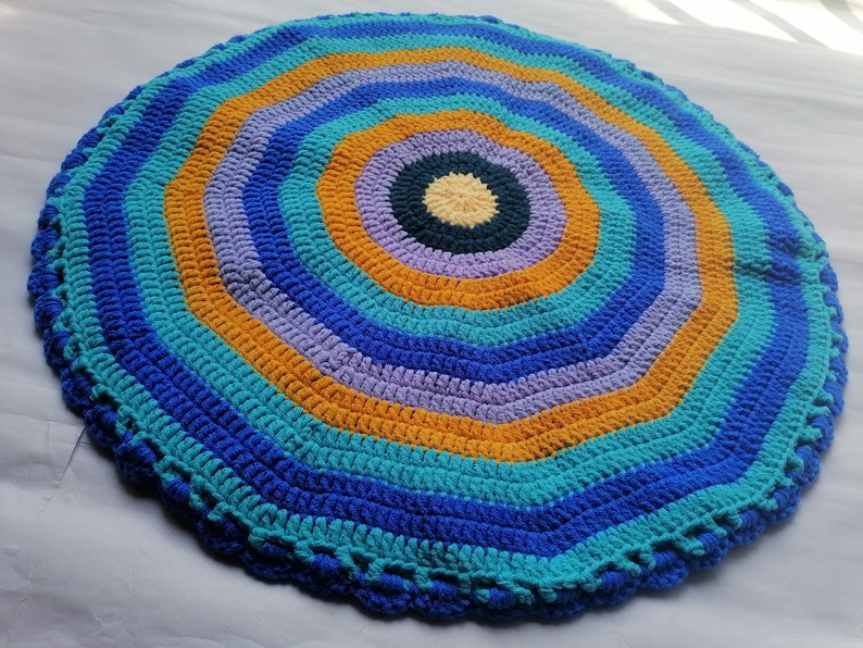 May include: A round crocheted rug with a multi-colored concentric circle pattern. The rug is made with blue, teal, purple, and orange yarn.