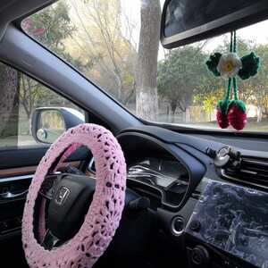 Puede incluir: Una funda de volante de crochet rosa con un patr&oacute;n blanco y negro. La funda est&aacute; instalada en un volante negro en el interior de un coche.