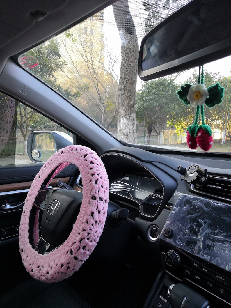 Puede incluir: Una funda de volante de crochet rosa con un patr&oacute;n blanco y negro. La funda est&aacute; instalada en un volante negro en el interior de un coche.