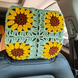 Puede incluir: Un reposacabezas de coche de ganchillo con fondo verde y cuatro girasoles amarillos con centros marrones.