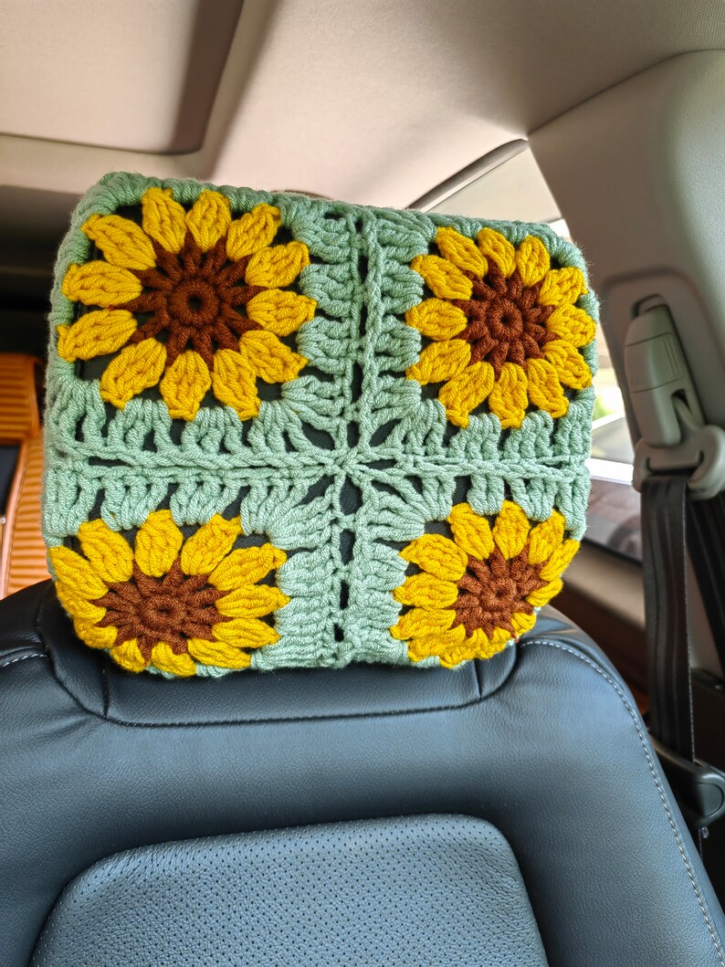 Puede incluir: Un reposacabezas de coche de ganchillo con fondo verde y cuatro girasoles amarillos con centros marrones.