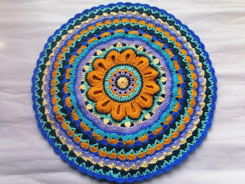 May include: A colorful crocheted mandala rug with a blue, yellow, orange, and purple design. The rug has a circular shape and a detailed pattern.
