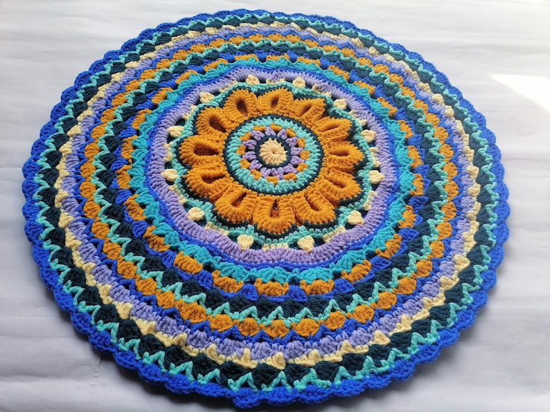 May include: A round crocheted rug with a colorful mandala design in shades of blue, yellow, and purple. The rug has a scalloped edge and is made with a variety of stitches.