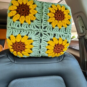 Puede incluir: Un reposacabezas de coche de ganchillo con forma de girasol. El reposacabezas est&aacute; hecho de hilo verde y amarillo y presenta cuatro girasoles.