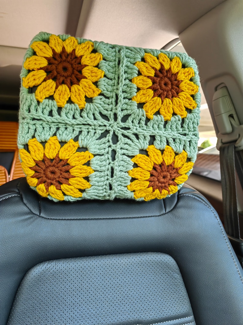 Puede incluir: Un reposacabezas de coche de ganchillo con forma de girasol. El reposacabezas est&aacute; hecho de hilo verde y amarillo y presenta cuatro girasoles.