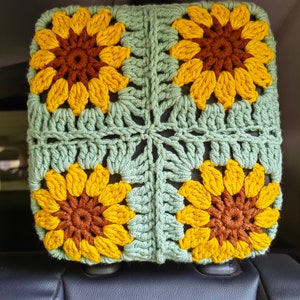 Puede incluir: Una funda para reposacabezas de asiento de coche de ganchillo con fondo verde y cuatro girasoles amarillos con centros marrones.