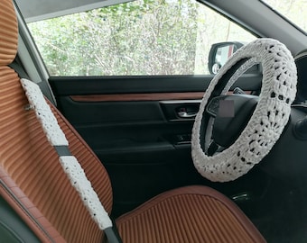 May include: White crocheted steering wheel cover in a car interior. The cover is made of a delicate, lacy pattern. The car seat is brown with a ribbed texture.