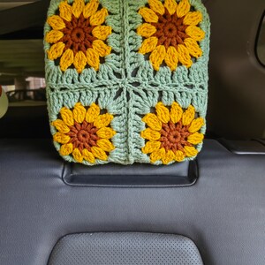 Puede incluir: Un reposacabezas de coche de ganchillo con forma de girasol. El reposacabezas est&aacute; hecho de hilo verde y amarillo y presenta cuatro girasoles de ganchillo.