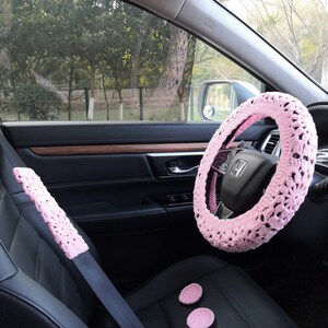 Puede incluir: Funda de volante de crochet rosa, funda de cintur&oacute;n de seguridad y dos posavasos de crochet rosa en el interior de un coche.