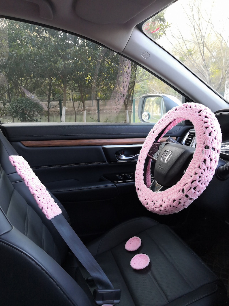 Puede incluir: Funda de volante de crochet rosa, funda de cintur&oacute;n de seguridad y dos posavasos de crochet rosa en el interior de un coche.