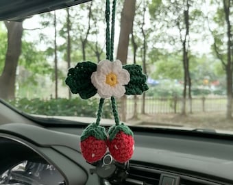May include: A crocheted car charm featuring a white flower with a yellow center and two red strawberries. The charm is hanging from the rearview mirror of a car.