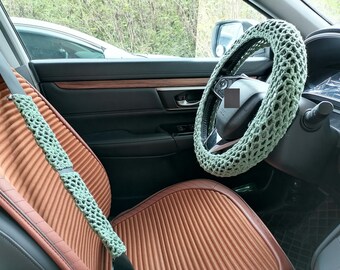 Op de afbeelding: Auto-interieur met een lichtgroene gehaakte stuurwielhoes en veiligheidsgordelcover. De autostoel is bruin met geribbelde details. Het interieur van de auto is zwart met houten afwerking.