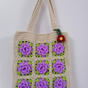 Puede incluir: Una bolsa de mano de crochet con fondo crema y flores moradas. La bolsa tiene dos asas largas y un pequeño adorno de flor roja.