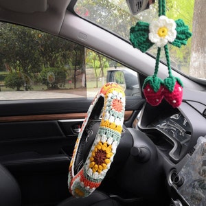 May include: A crocheted steering wheel cover with a sunflower design in shades of yellow, orange, and brown. The cover is white with green and orange accents. A crocheted flower and strawberry charm hangs from the rearview mirror.