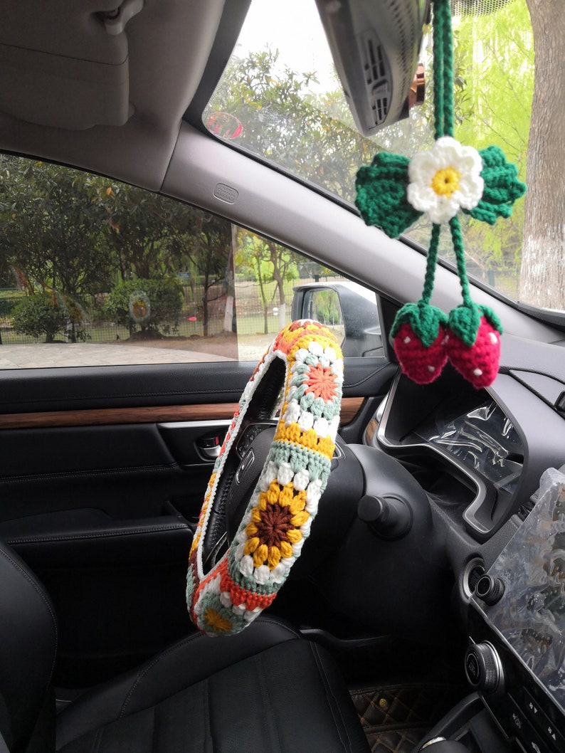 May include: A crocheted steering wheel cover with a sunflower design in shades of yellow, orange, and brown. The cover is white with green and orange accents. A crocheted flower and strawberry charm hangs from the rearview mirror.