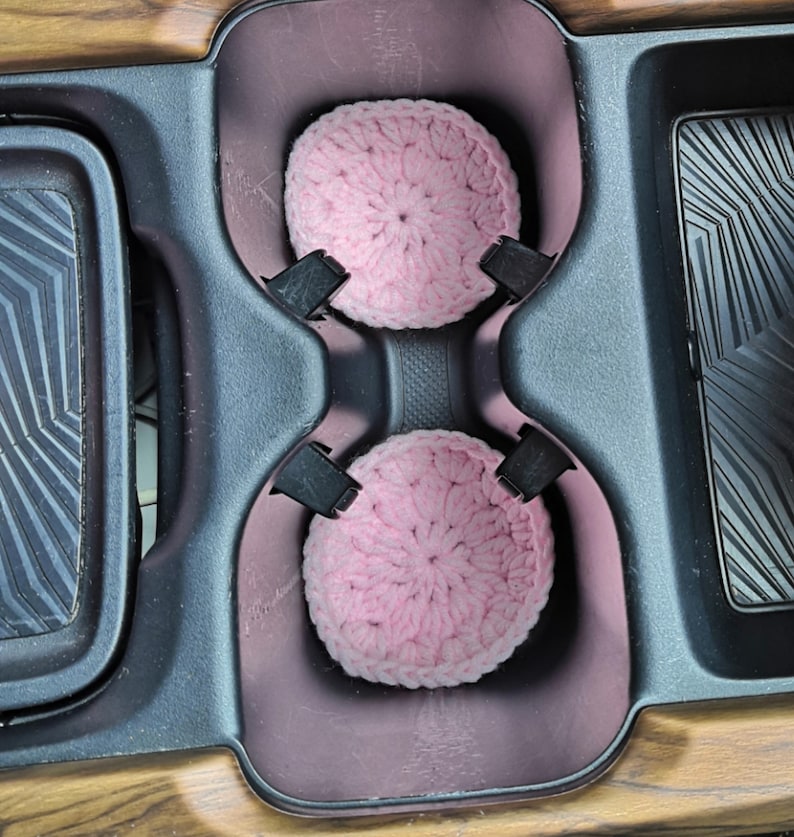 Puede incluir: Dos posavasos de ganchillo de color rosa en el portavasos de un coche.