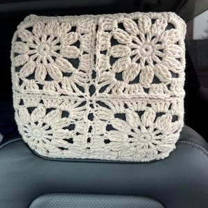 May include: A cream-coloured, crocheted headrest cover with a floral pattern. The cover is made from soft, textured yarn and sits on a black car seat headrest. The crochet design creates a dimensional, decorative effect.