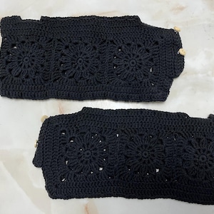 Könnte beinhalten: Zwei schwarze Armstulpen aus Häkelarbeit mit floralem Muster. Die Armstulpen sind aus dickem Garn gefertigt und haben einen Knopfverschluss. Das Häkeldesign zeigt ein sich wiederholendes Blumenmotiv und verleiht einen handgemachten Charme.